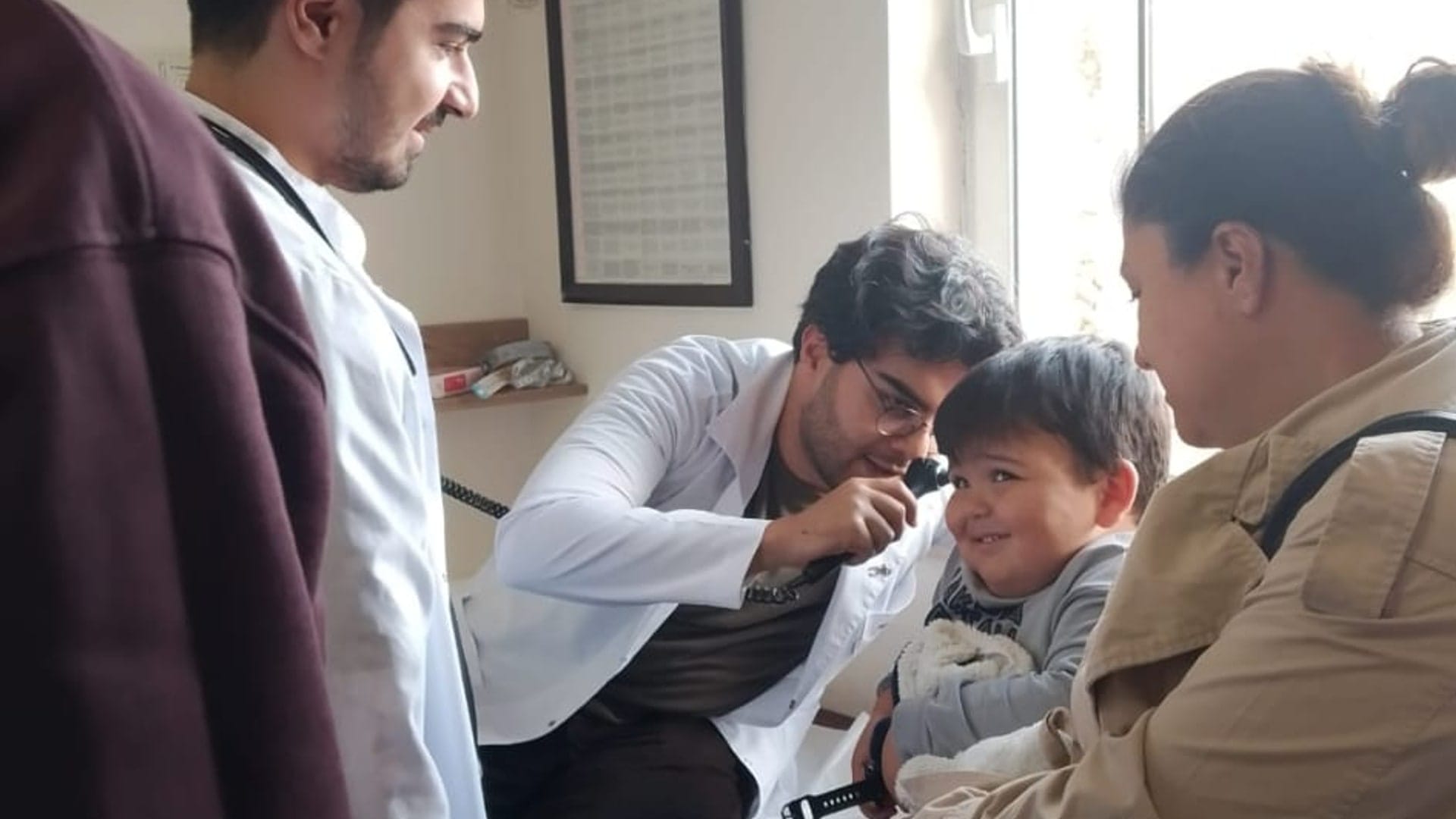 A physician examining a young child's ear with an otoscope, with the child's mother present and a colleague nearby, in a clinic exam room.