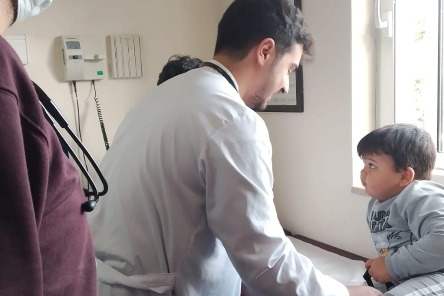 A physician attending to a toddler seated on an exam table, with a colleague present.